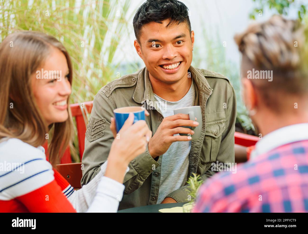 Fröhlicher Mann und Frau aus verschiedenen Rassen mit einer Tasse Kaffee, die lächelt und Freunden zuhört, während sie im Café im Freien am Tisch sitzen Stockfoto