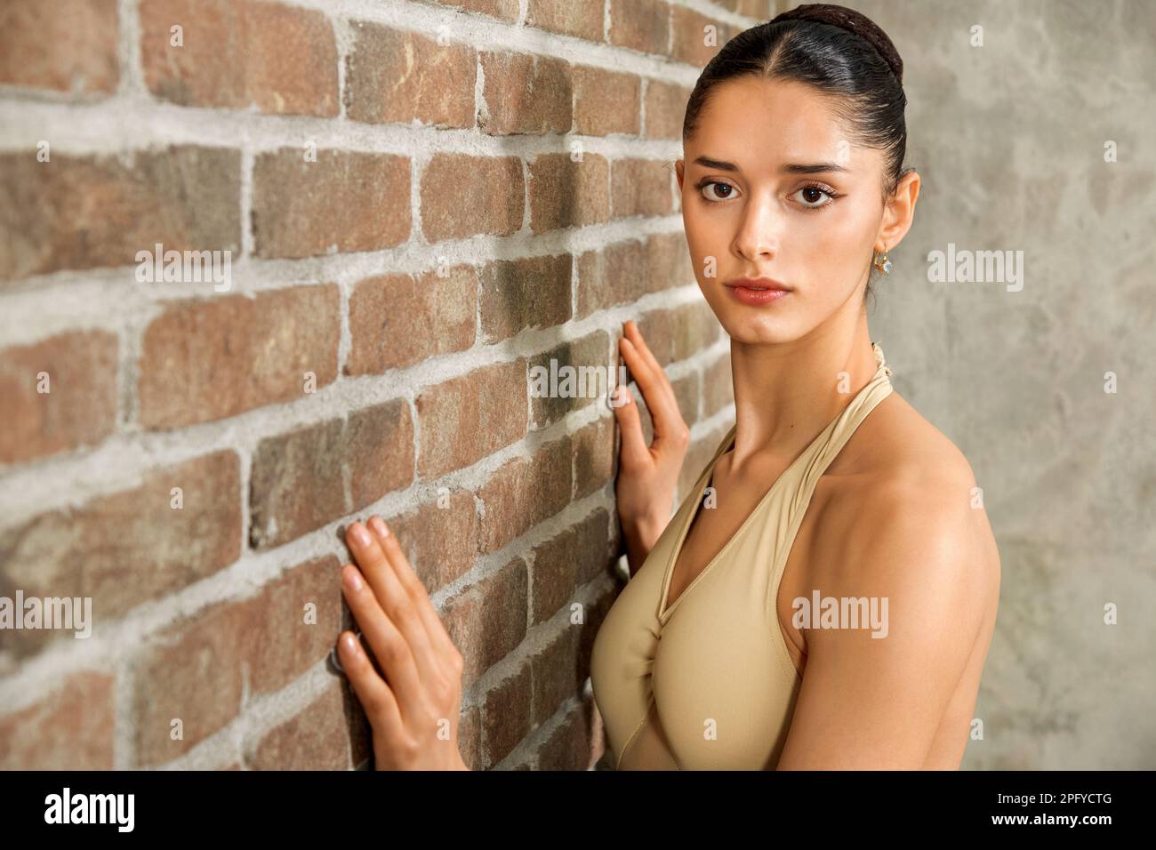 Attraktive junge Balletttänzerin mit Brötchenhaarschnitt, die in einem modernen, hellen Studio neben einer Backsteinwand vor die Kamera schaut Stockfoto