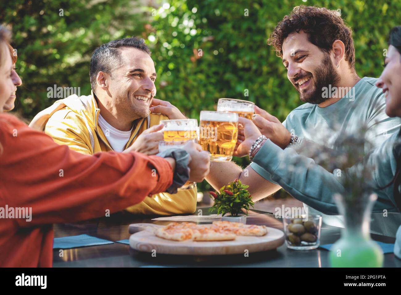 Eine Gruppe von fünf Freunden, die an einem Tisch in einem Garten sitzen und Bierbecher zum Anstoßen aufbringen. Eine köstliche Pizza liegt auf einem Schneidebrett vor ihnen - Fokus o Stockfoto