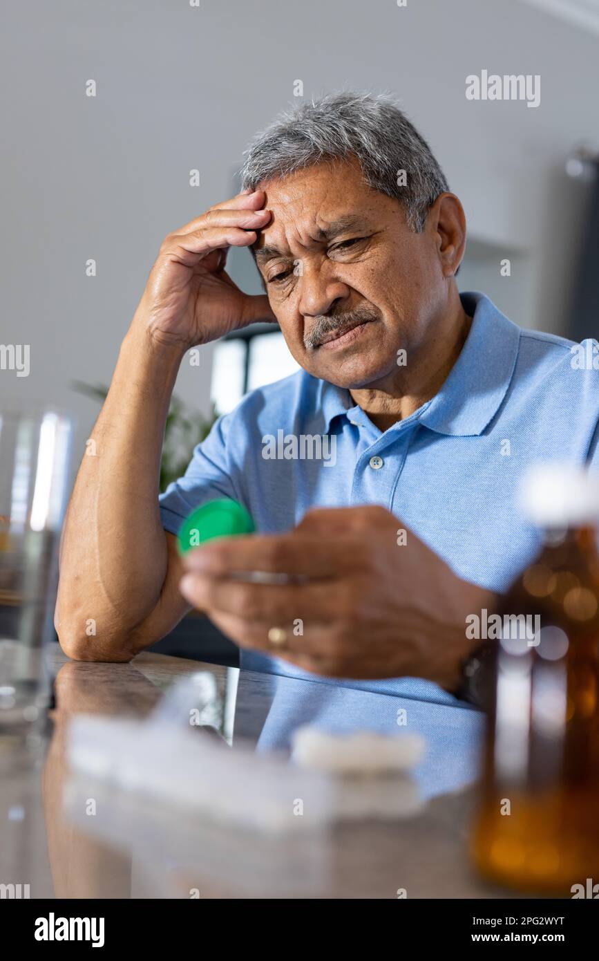 Nahaufnahme eines traurigen, birassischen Seniorenmannes mit Kopf in der Hand, der zu Hause auf dem Tisch eine verschriebene Flasche anschaut Stockfoto