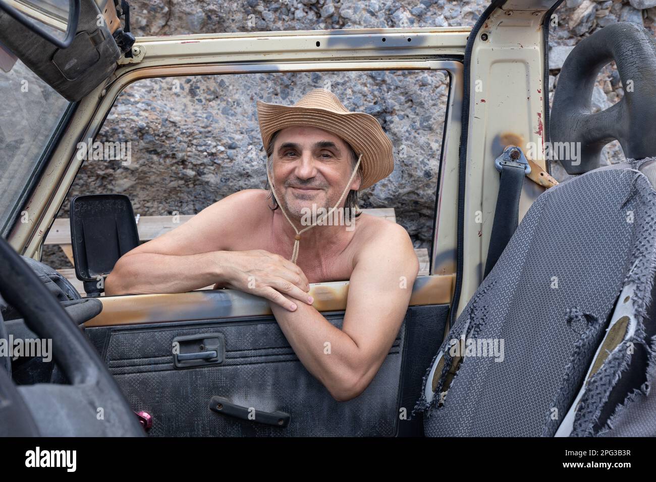 Ein erwachsener Mann mit Hut lehnt sich an ein altes Auto und posiert in einem Fenster, Griechenland, Kreta Stockfoto