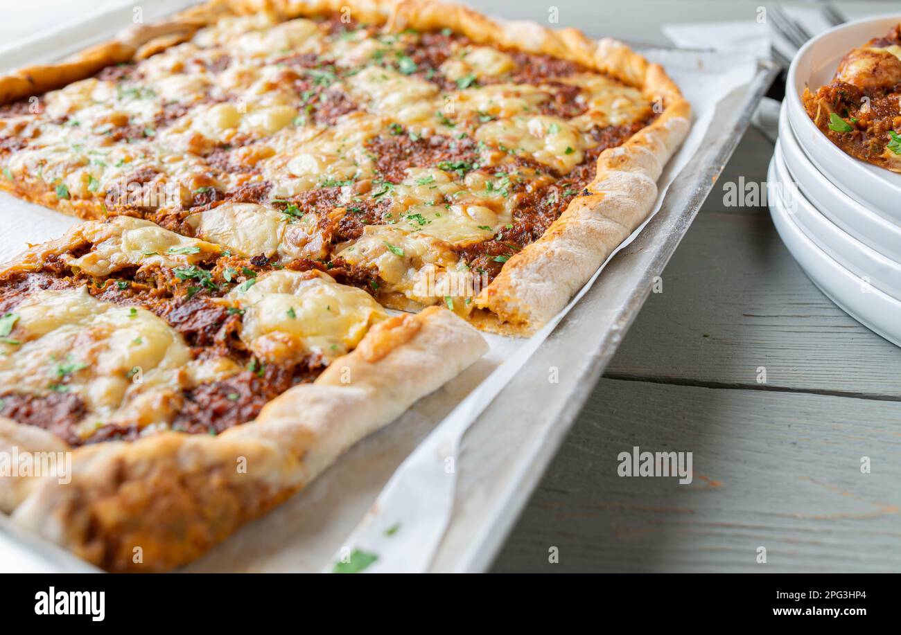 Quadratische Pizza mit Hackfleisch, Käse, Tomaten, Paprika, Zwiebeln und Kräutern auf einem Backblech Stockfoto