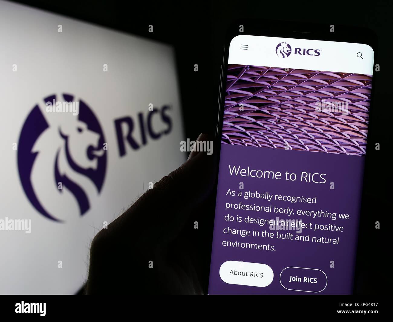 Person mit Mobiltelefon und Website der Royal Institution of Chartered Surveyors (RICS) auf dem Bildschirm mit Logo. Konzentrieren Sie sich auf die Mitte des Telefondisplays. Stockfoto