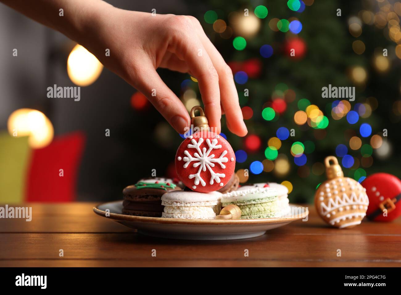 Eine Frau, die Weihnachtsmakarons vom Teller am Holztisch nimmt, vor verschwommenen festlichen Lichtern, Nahaufnahme Stockfoto