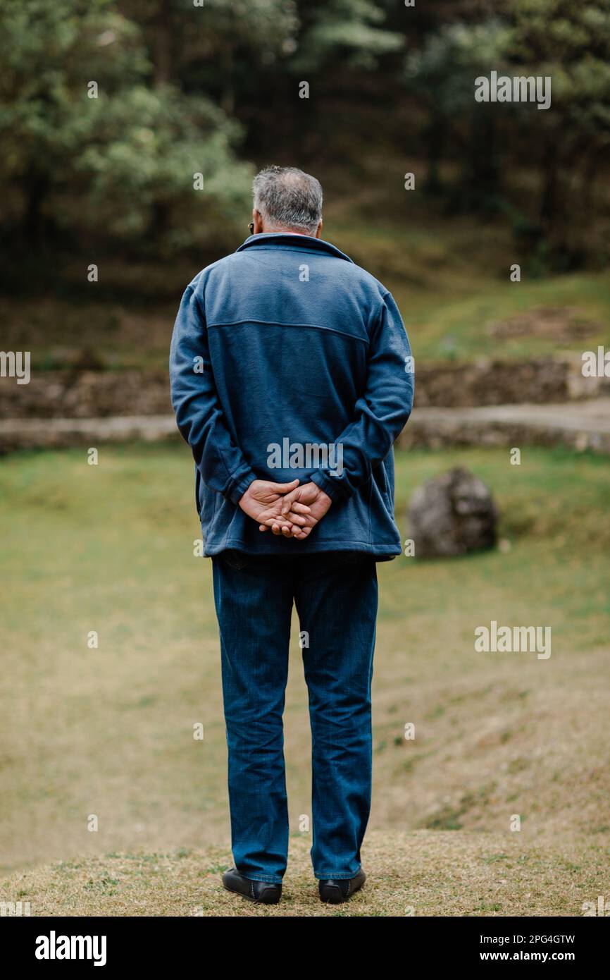 Ein älterer Mann, der auf einer Wiese steht, mit den Händen auf dem Rücken und in die Ferne blickt Stockfoto