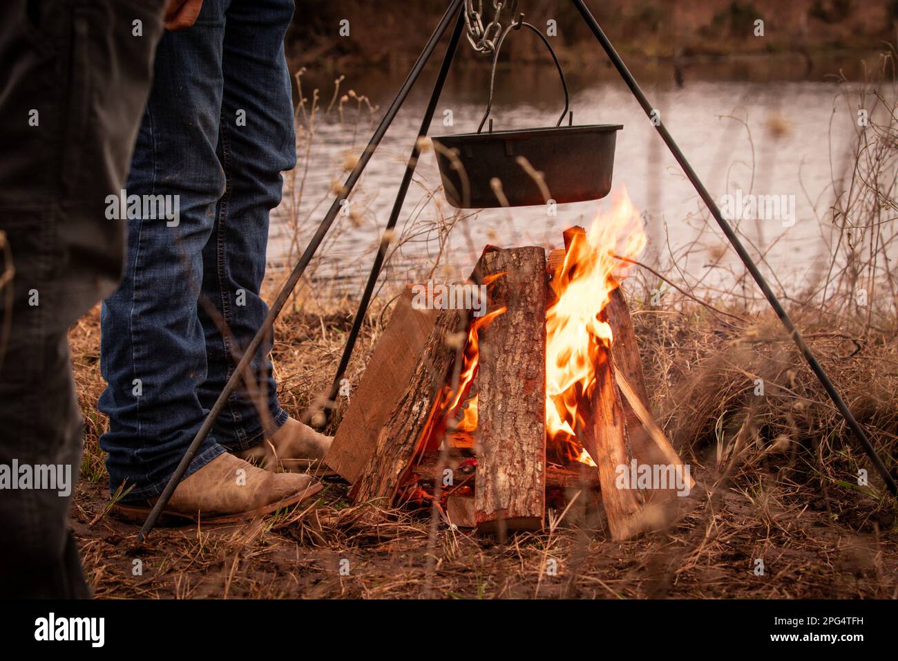 2 Cowboys kochen am Lagerfeuer Chili in einem Eisentopf neben dem See. Stockfoto