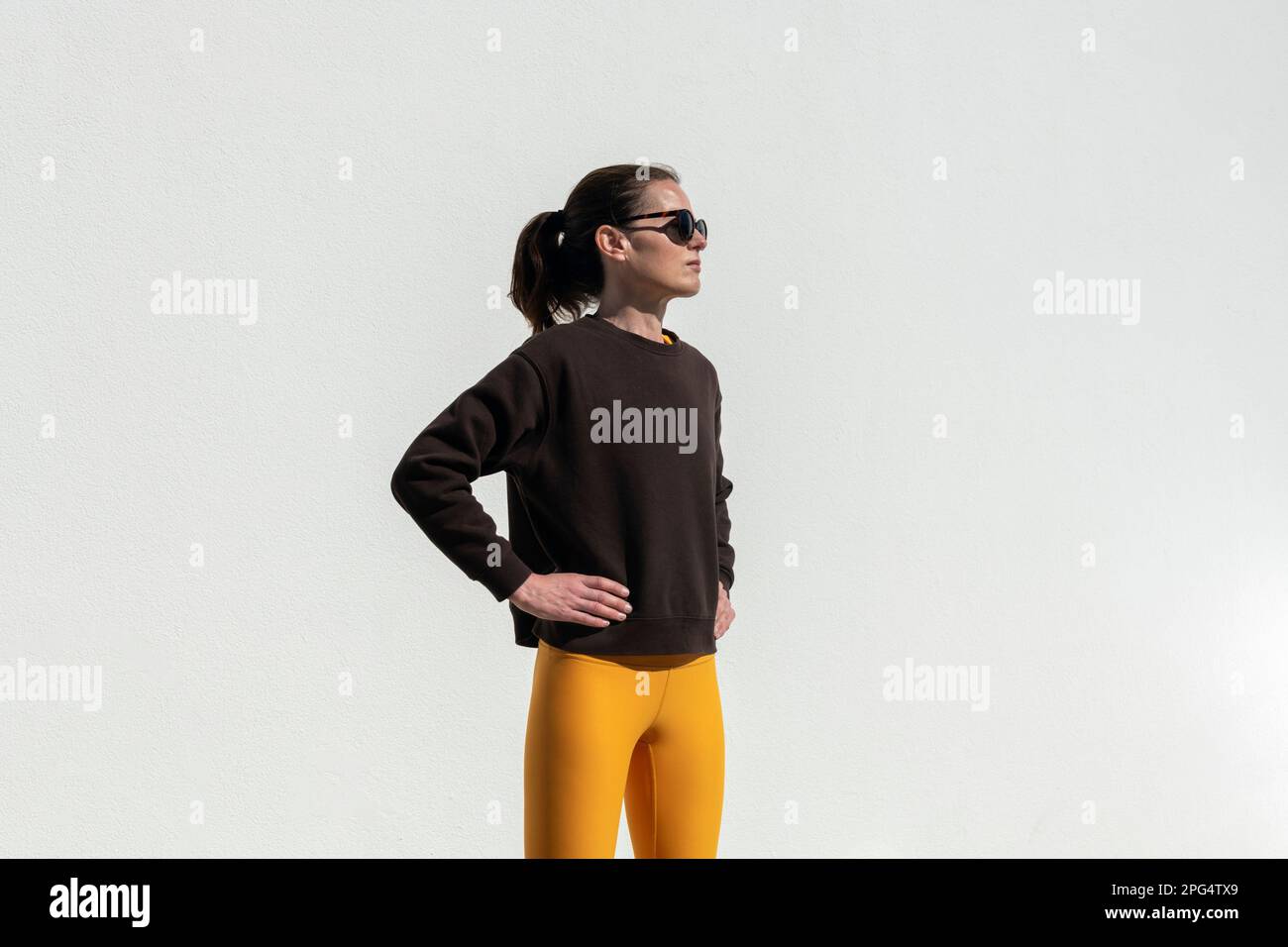 Fit sportliche Frau mit Sweatshirt und Yoga-Hose, die an einer weißen Wand steht. Stockfoto