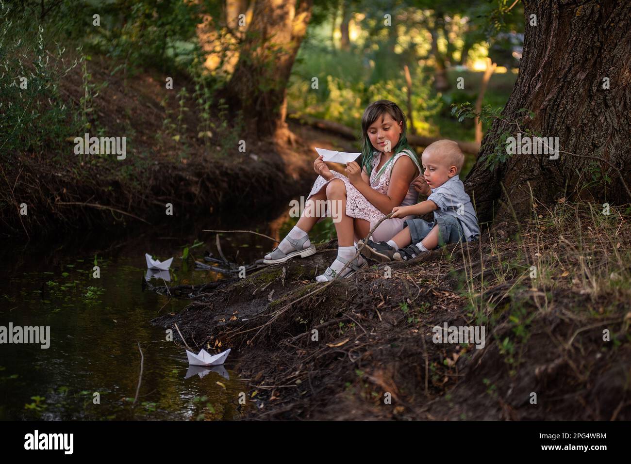 Mädchen und Junge sitzen am Ufer des Flusses und werfen das Origami-Boot aus weißem Papier ins Wasser. Eine Teenager-Schwester mit grünem Haar umarmt den kleinen Bruder. Stockfoto