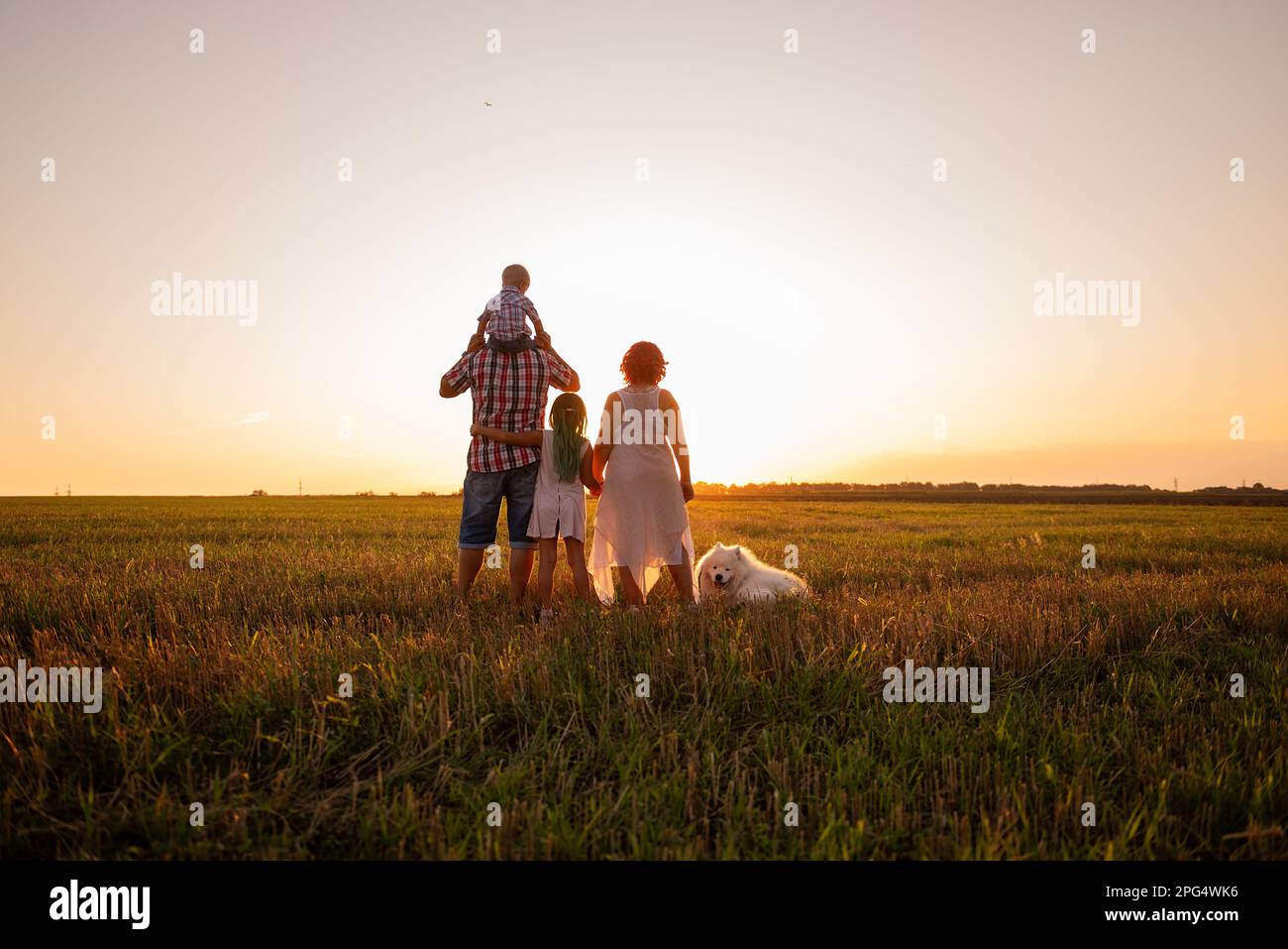 Silhouette der Familie mit Samoyerten Hund bei Sonnenuntergang. Gesichtslos, Leute stehen mit dem Rücken zur Kamera. Das Konzept des Reisens, der Freiheit, des Vertrauens in die Fut Stockfoto