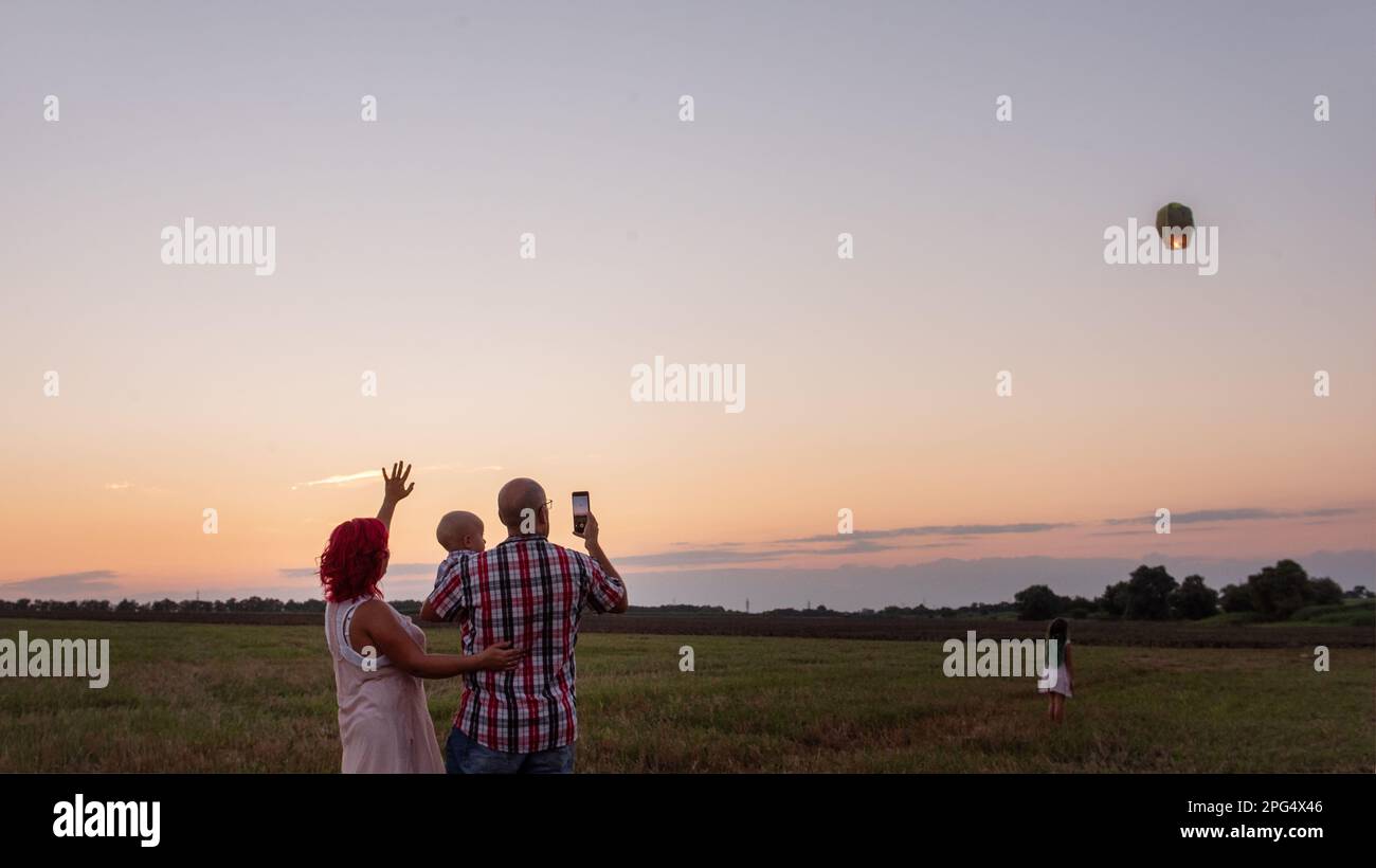 Diversity Family fotografiert mit der gelben Himmelslaterne des Smartphones im Feld bei Sonnenuntergang. Mutter Vater hält den kleinen Sohn, Tochter winkt mit den Händen. Ich mache wis Stockfoto