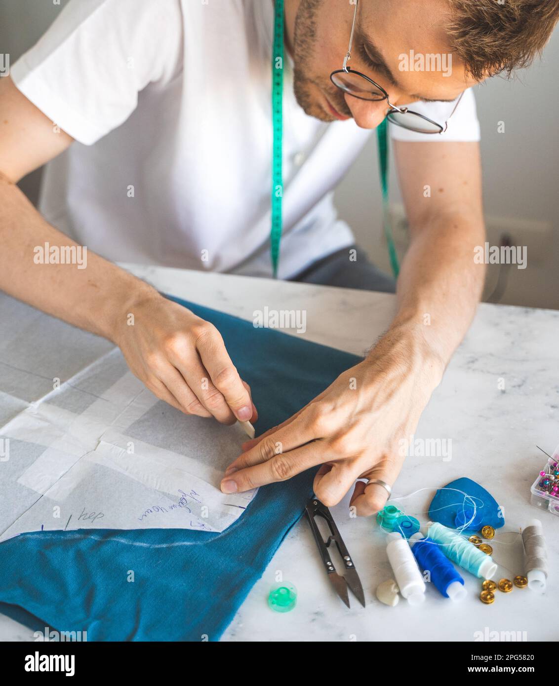 Ein männlicher Heimwerker überträgt eine Vorlage in ein Stoffstudio. Ein selbstlernender Schneider in einem weißen T-Shirt und einer Brille arbeitet mit Stoff und Muster Stockfoto