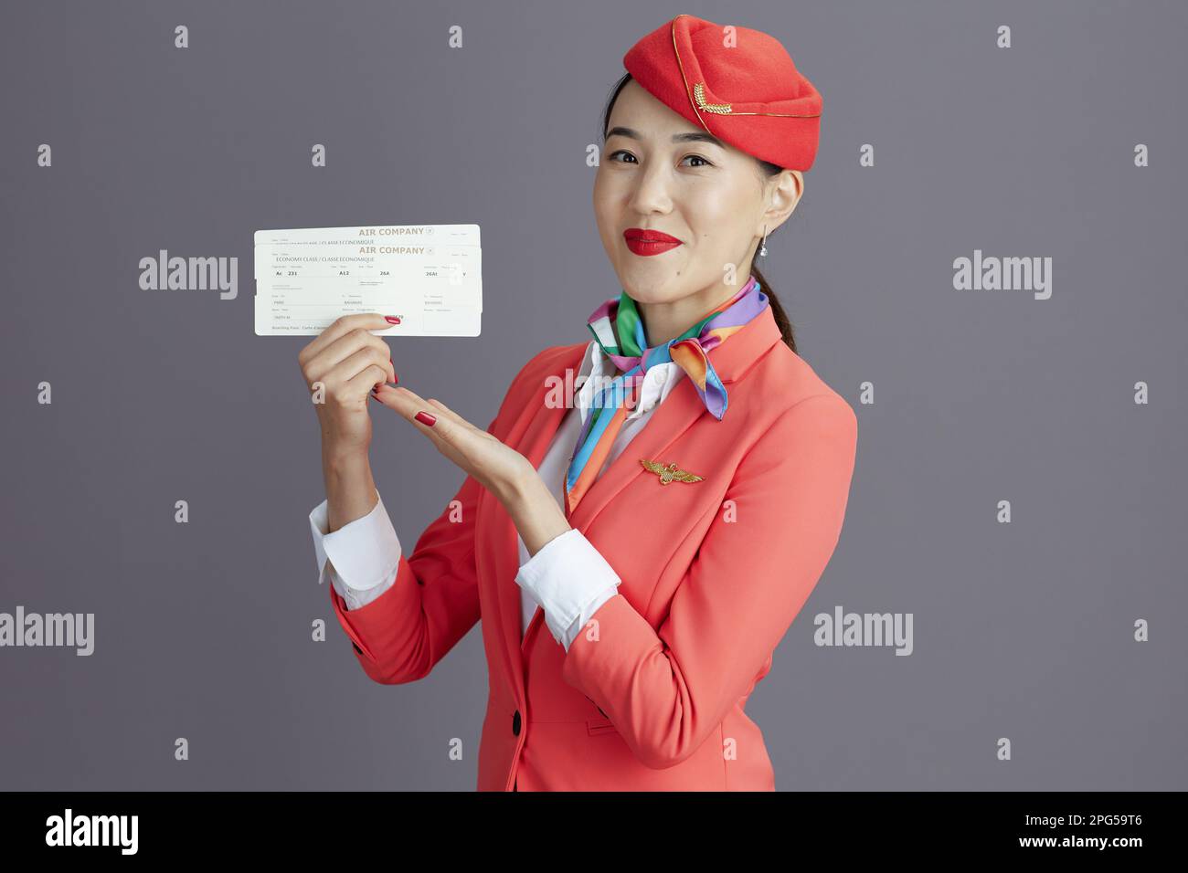 Fröhliche, elegante Stewardess Asiatin in rotem Rock, Jacke und Hut Uniform mit Flugtickets vor grauem Hintergrund. Stockfoto