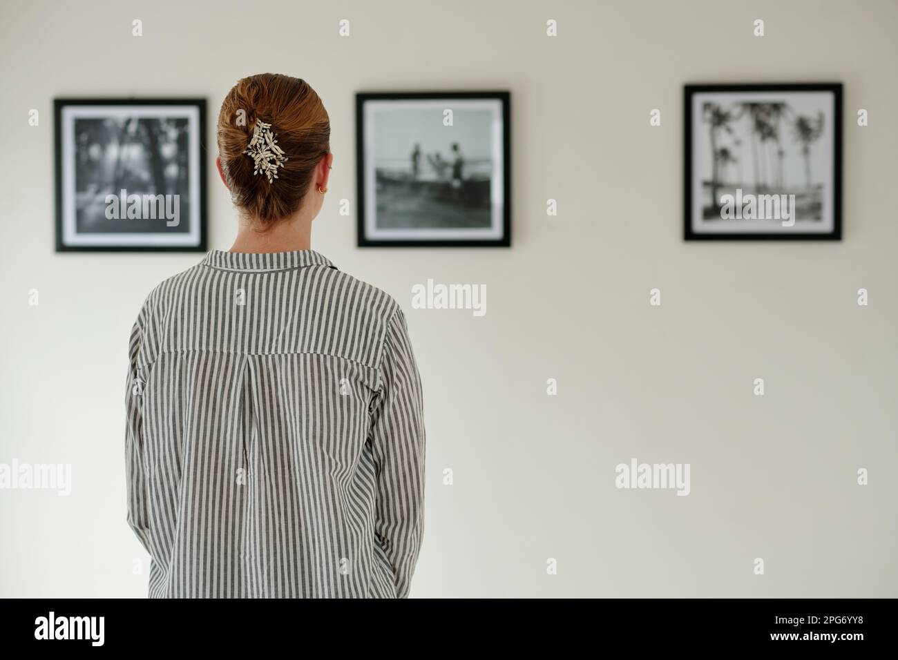 Rückansicht einer jungen Frau in gestreiftem Hemd, die vor der Wand steht, mit Bildern oder Kunstwerken in schwarzen Rahmen, während sie die Kunstgalerie besucht Stockfoto