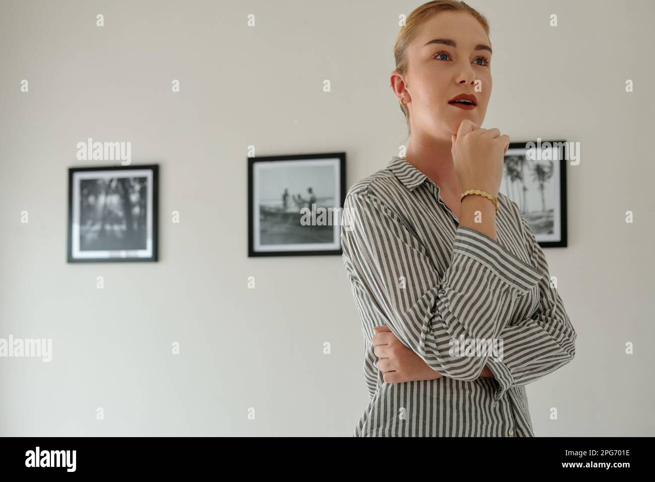 Junge Frau in Freizeitkleidung, die sich Kunstwerke mit Aufmerksamkeit ansieht, während sie vor der Kamera mit einer Reihe von Bildern an der Wand steht Stockfoto
