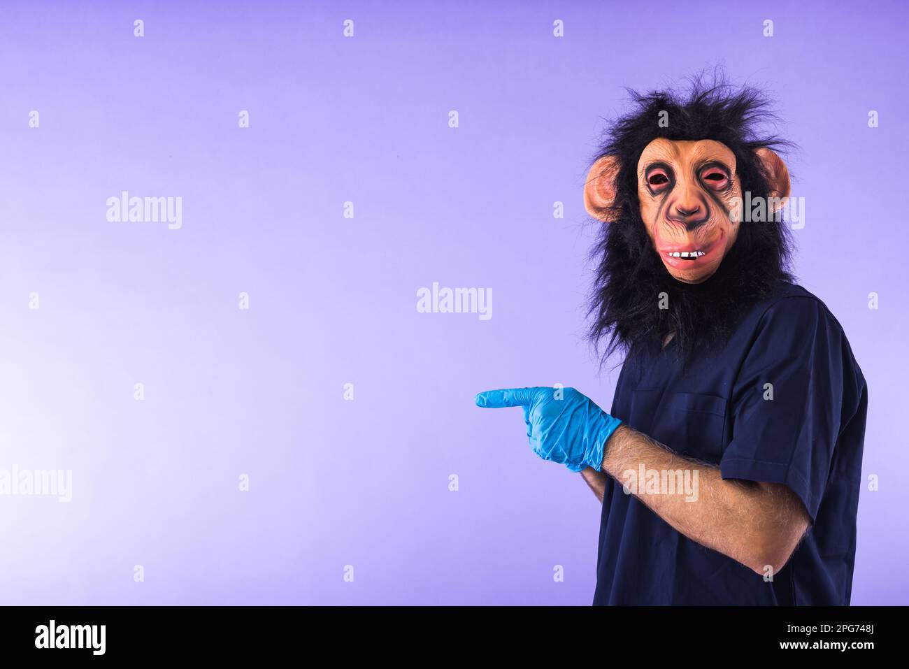 Verkleidete Person in einer Affenmaske und Arztkostüm, die mit den Fingern zur Seite zeigt, auf lila Hintergrund. AFFENPOCKEN, Pandemie, Virus, Epidemie, Stockfoto