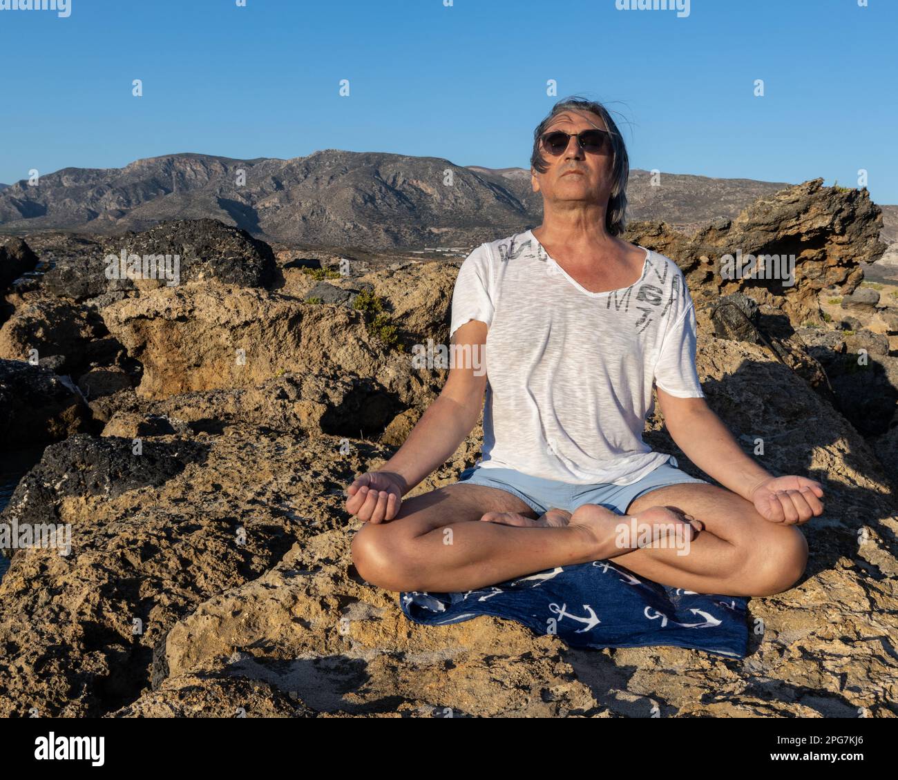 Ein grauhaariger Mann bei Sonnenuntergang macht Yoga vor dem Hintergrund einer felsigen Küste und Berge, Griechenland, Kreta Stockfoto