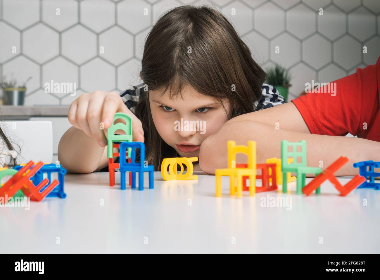 Das kleine Mädchen baut mit seinen Käfern einen kleinen grünen Stuhl und hält ihn in der Nähe vieler farbenfroher Spielzeuge auf einem weißen Tisch. Stuhlleiter Stockfoto