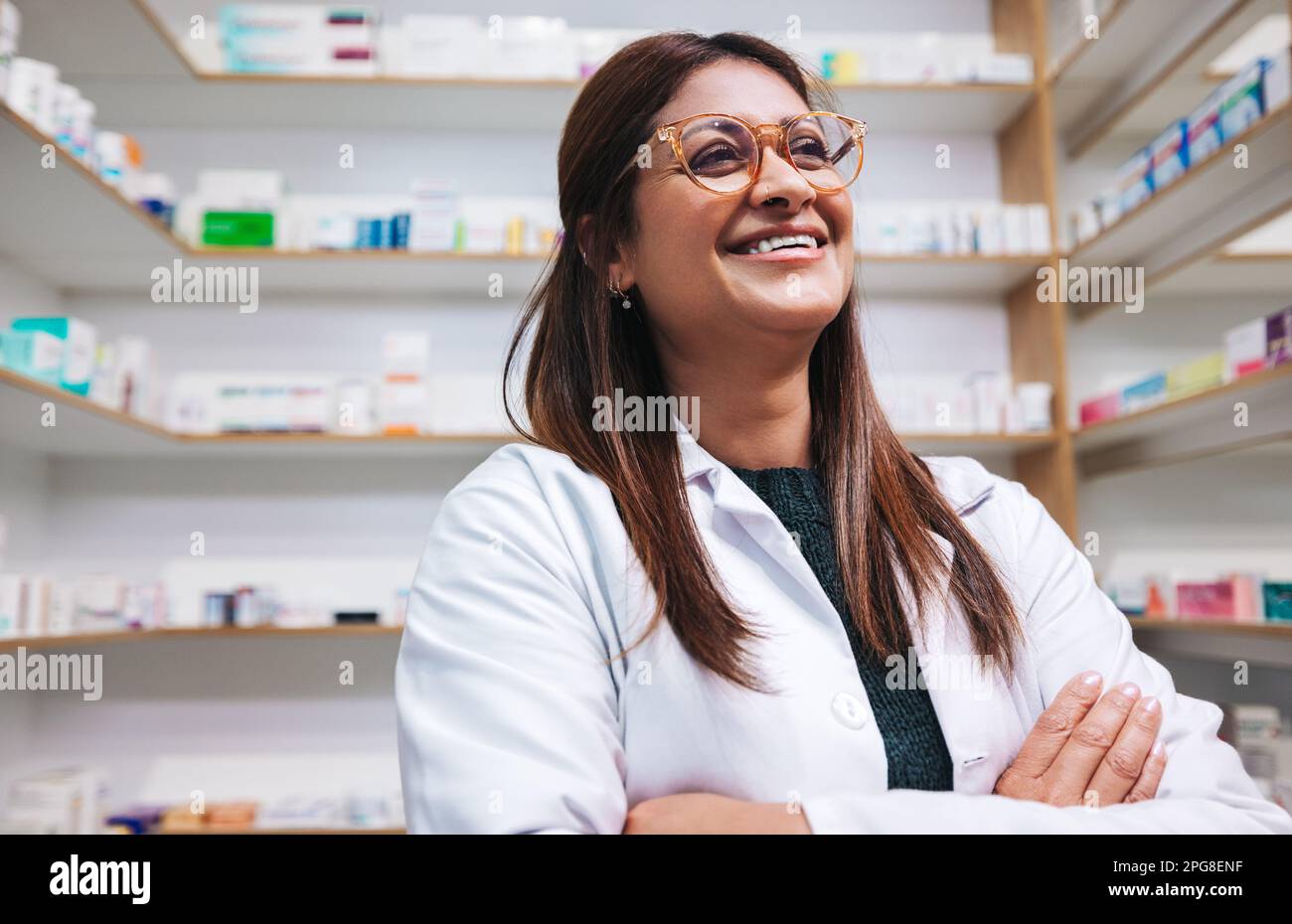 Glückliche Apothekerin, die in einer Apotheke mit gekreuzten Armen steht. Reife Frau, die in einer Apotheke arbeitet. Gesundheitshelferin in einer Apotheke. Stockfoto