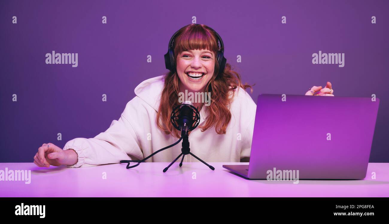 Glückliche junge Frau, die lacht, während sie eine Live-Radiosendung in einem Studio aufnimmt. Fröhliche junge Frau, die eine Audioübertragung in Neonlila moderiert. Jung Stockfoto