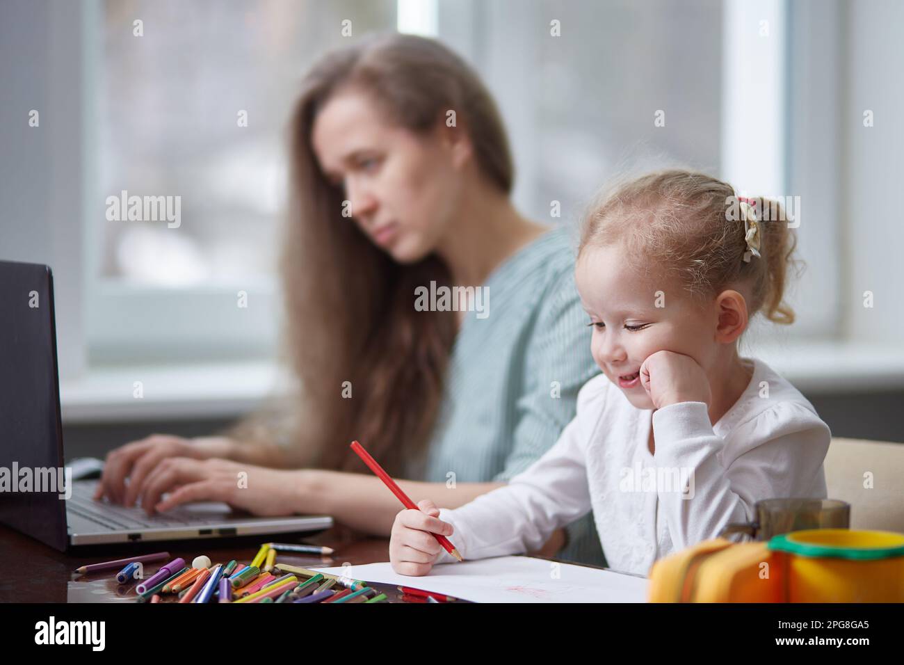 Eine freiberufliche Frau, die zu Hause mit einem Kind arbeitet. Konzentrier dich auf das Kind. Das Mädchen malt mit Bleistiften, die Mutter arbeitet. Freiberuflich, Muttertagskonzept. Stockfoto