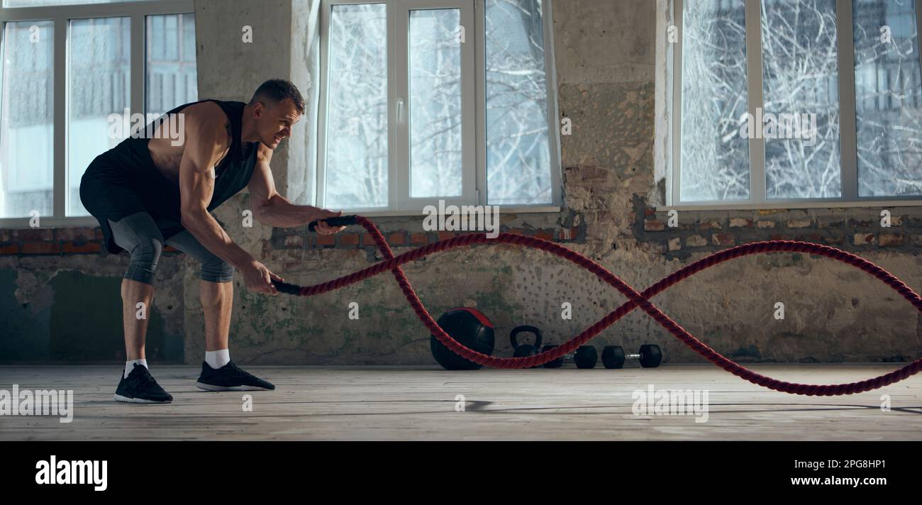 Konzentrierter junger Mann mit sportlichem, muskulärem, kräftigem Körpertraining und Kampfseilen im Innenbereich. Workout. Konzept des sportlichen Lebensstils Stockfoto