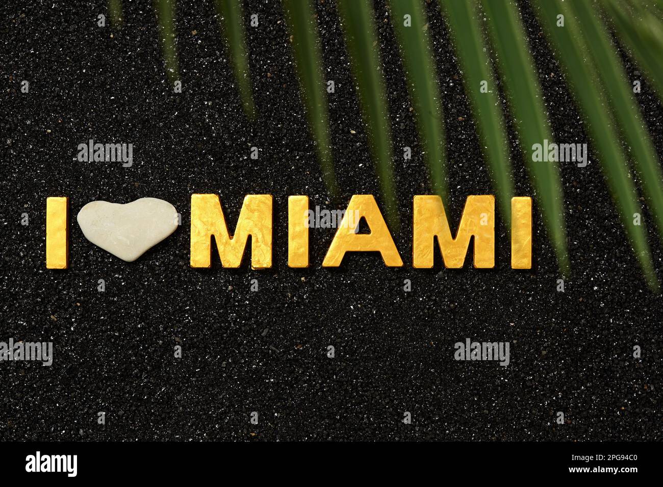 I Love Miami Beach - Goldene Buchstaben in schwarzem Sand mit Palmenblättern darüber Stockfoto