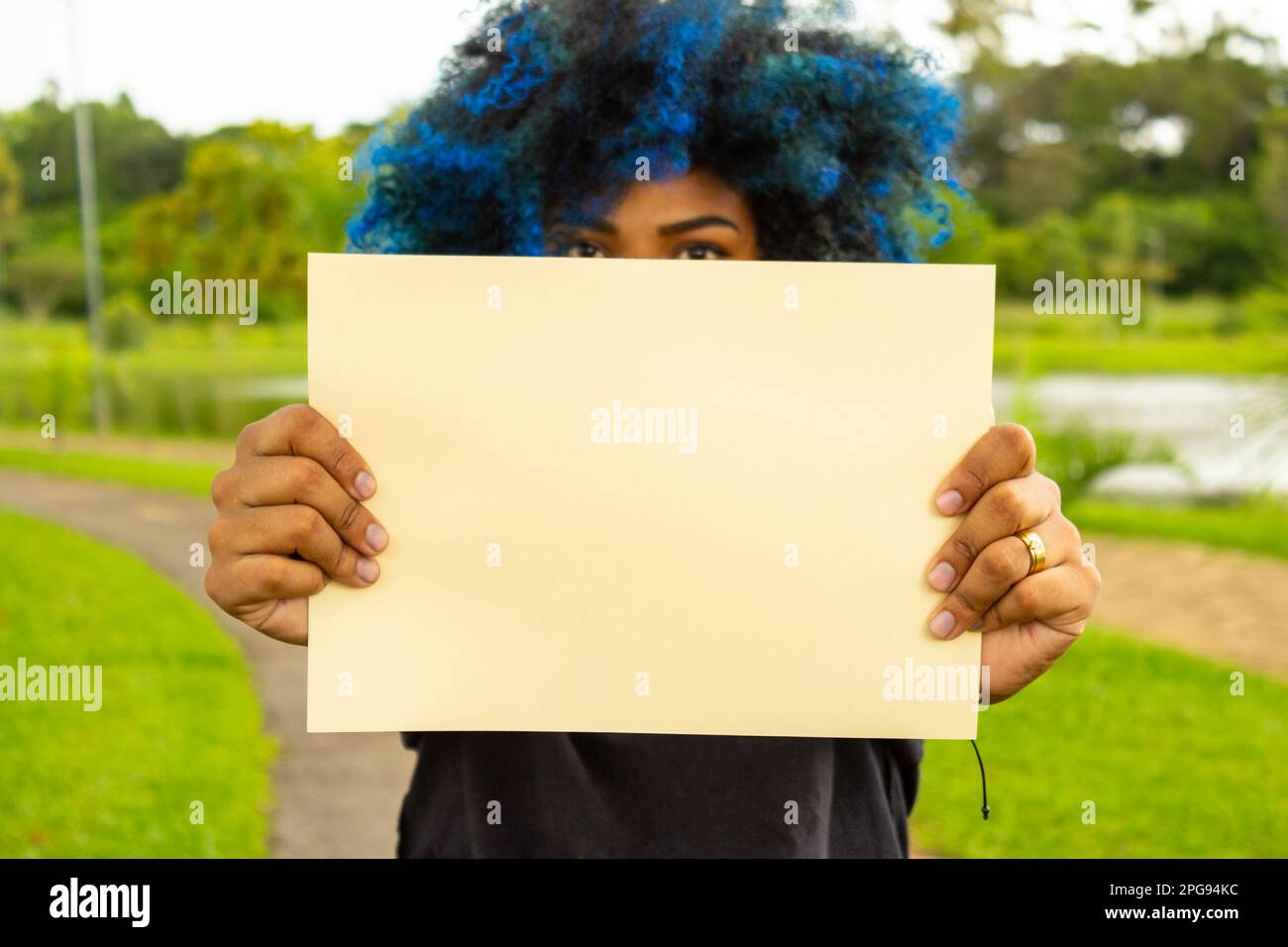 Goiania, Goias, Brasilien – 21. März 2023: Eine junge Frau mit blau gefärbtem Haar, hinter einem leeren Poster verborgenem Gesicht und einer Landschaft im Hintergrund Stockfoto