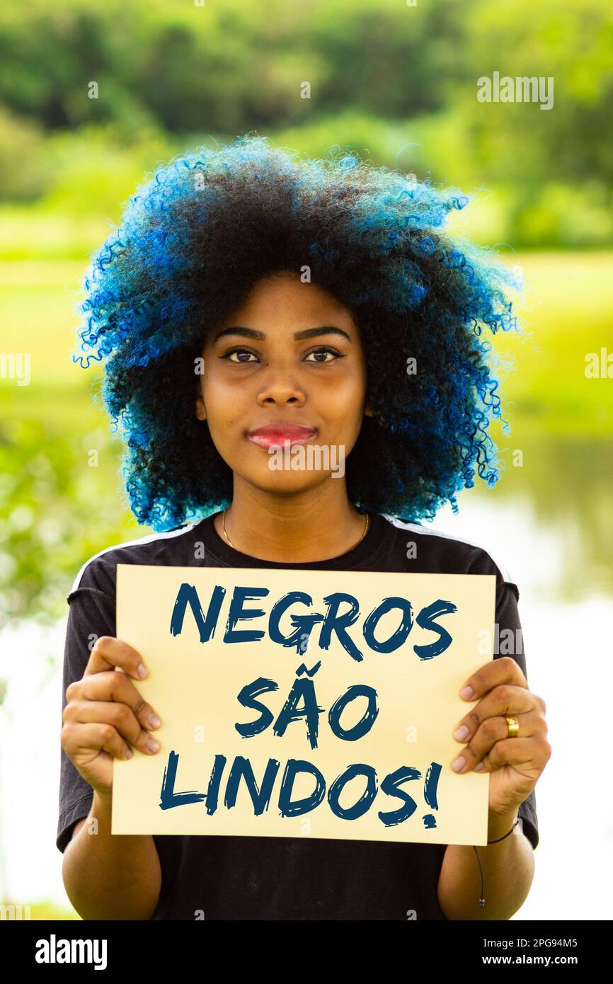 Goiania, Goias, Brasilien – 21. März 2023: Eine junge Frau mit gefärbtem blauem Haar, die ein Schild mit dem Text "Schwarze sind wunderschön" in der Hand hält. Stockfoto