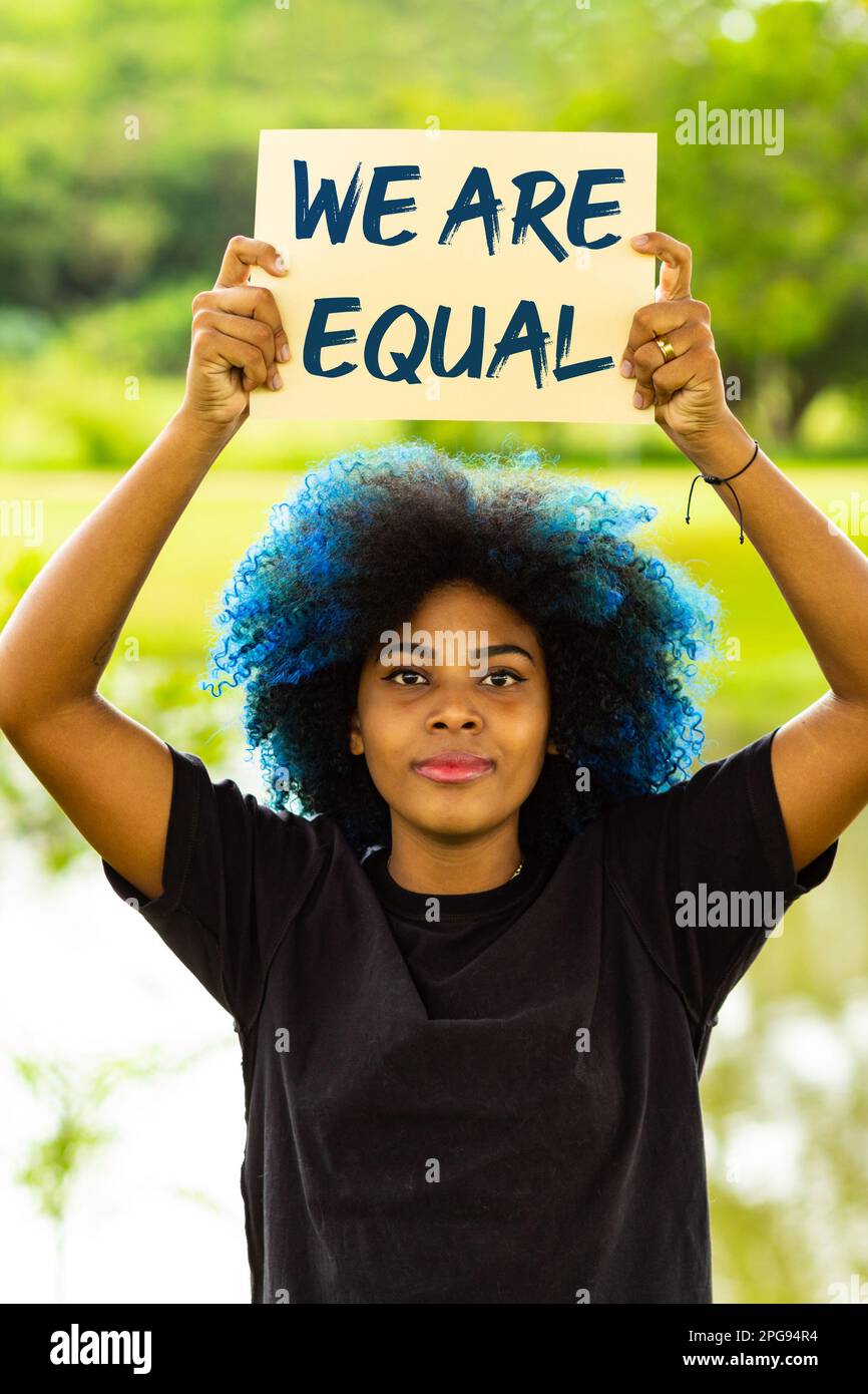 Goiania, Goias, Brasilien – 21. März 2023: Eine junge Frau mit gefärbtem blauem Haar, die ein Schild mit dem Text "We are Equal" in der Hand hält, mit einer Landschaft im Bac Stockfoto