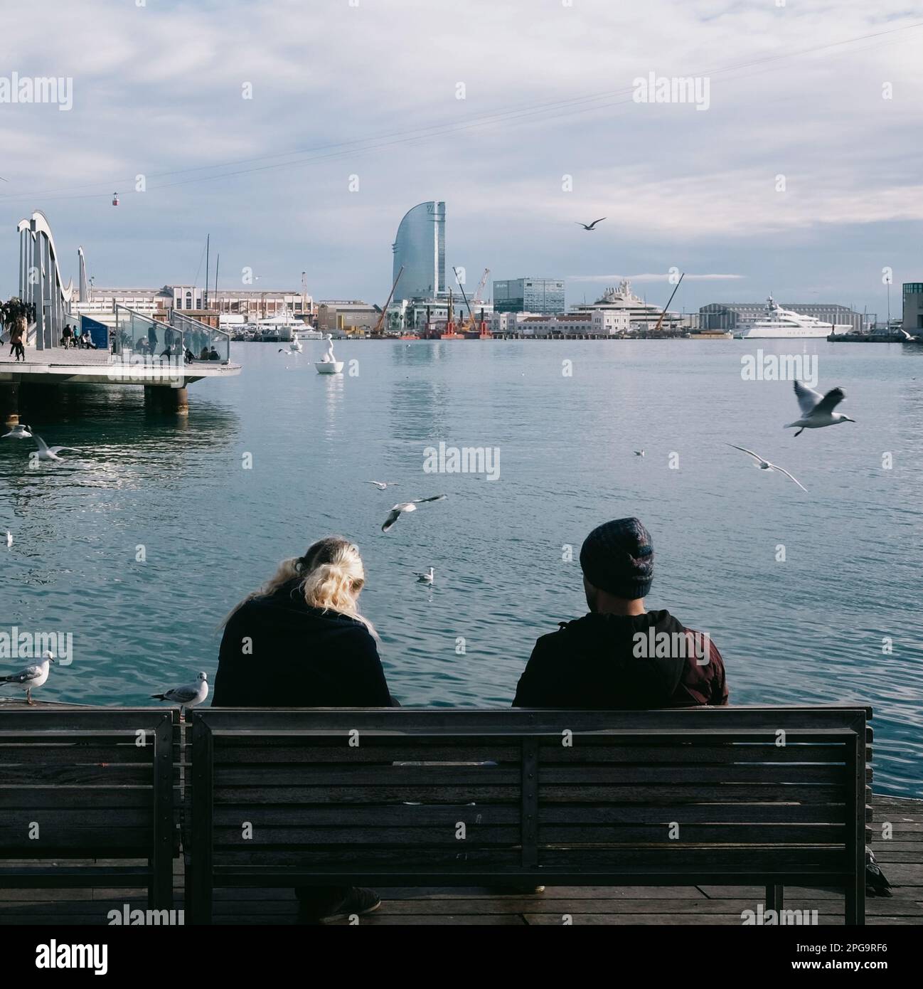 Ein Mann und eine Frau sitzen auf einer Bank und genießen den Blick auf die hoch aufragenden Vögel über dem Meer in Barcelona. Stockfoto