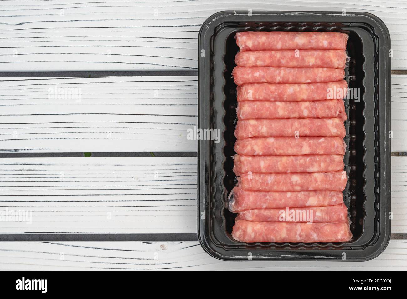 Rohe Frühstückswürste mit Hühnern in schwarzem Kunststoffbehälter, Nahaufnahme auf weißem Holzbrett, flach liegend mit Kopierbereich Stockfoto