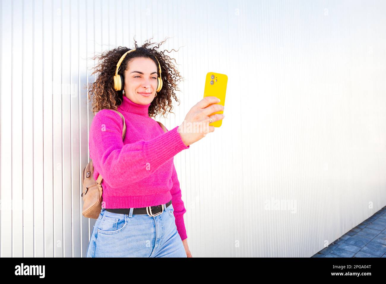 Junge weiße Frau mit lockigem Haar nimmt ein Selbstporträt mit ihrem Smartphone auf, während sie sich an die Wand lehnt und Musik über Kopfhörer hört. Stockfoto