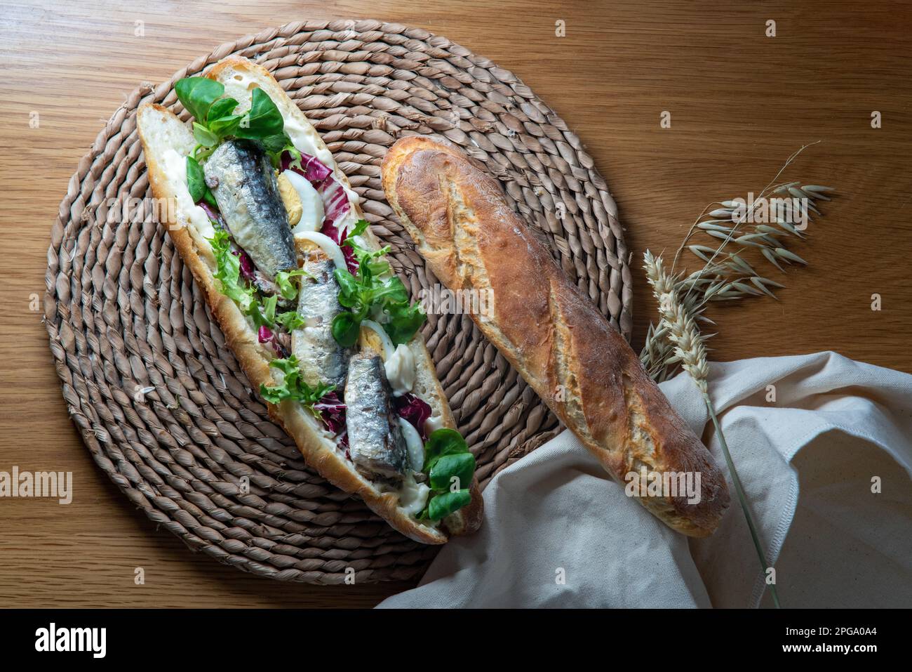 Baguette-Sandwich mit Sardine, Eiern, gemischtem Salat und Mayonnaise, weit geschnitten, serviert auf einer rustikalen Unterlage, auf einem Holztisch mit Backtuch, Stockfoto