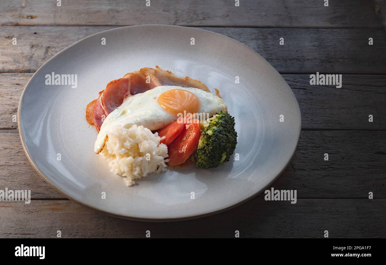 Eierfrühstück. Eier auf geröstetem Schinken mit Reis und gedämpftem Gemüse, Karotten und Brokkoli, serviert auf einem Teller auf einem Holztisch. Stockfoto