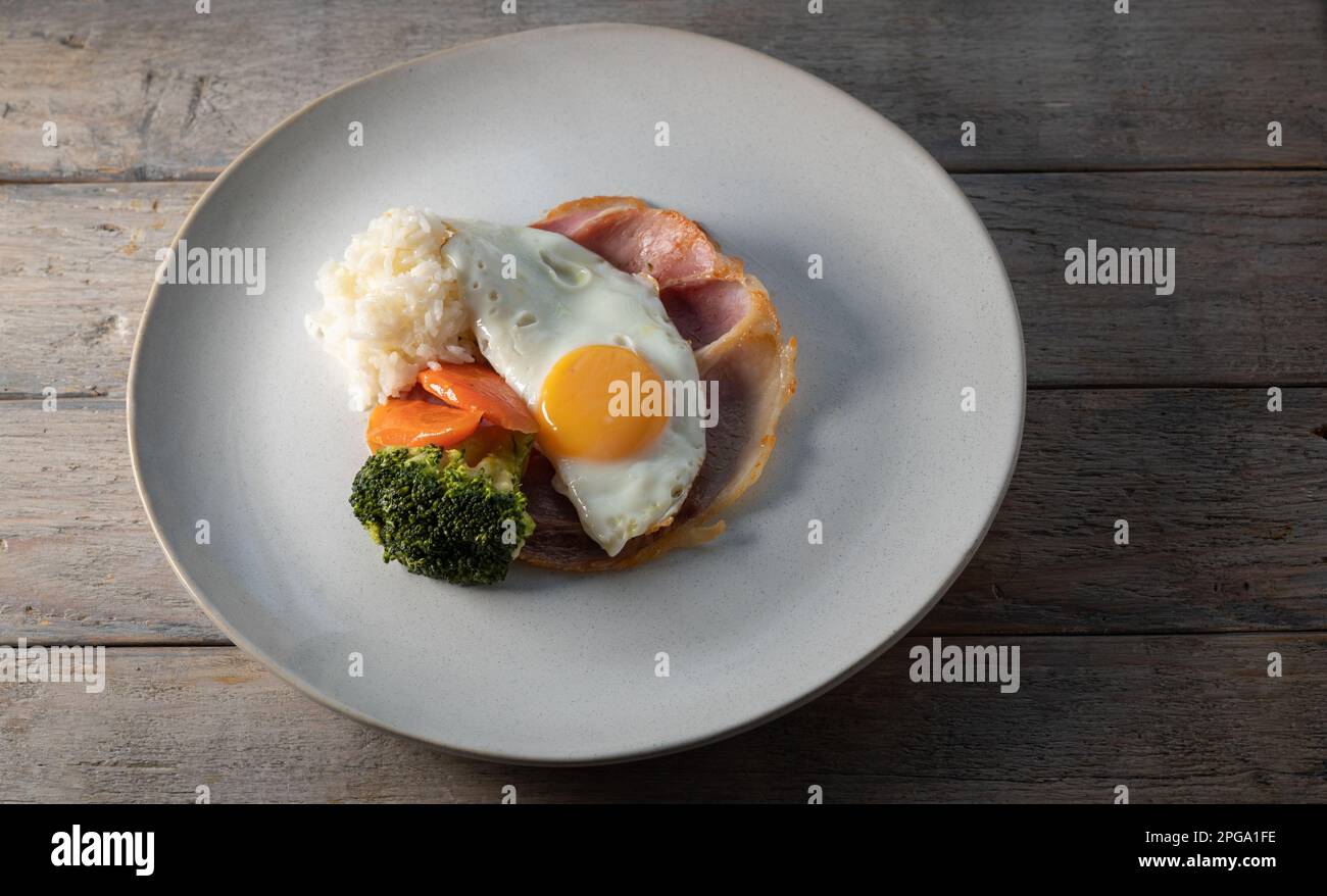 Eierfrühstück. Eier auf geröstetem Schinken mit Reis und gedämpftem Gemüse, Karotten und Brokkoli, serviert auf einem Teller auf einem Holztisch. Stockfoto