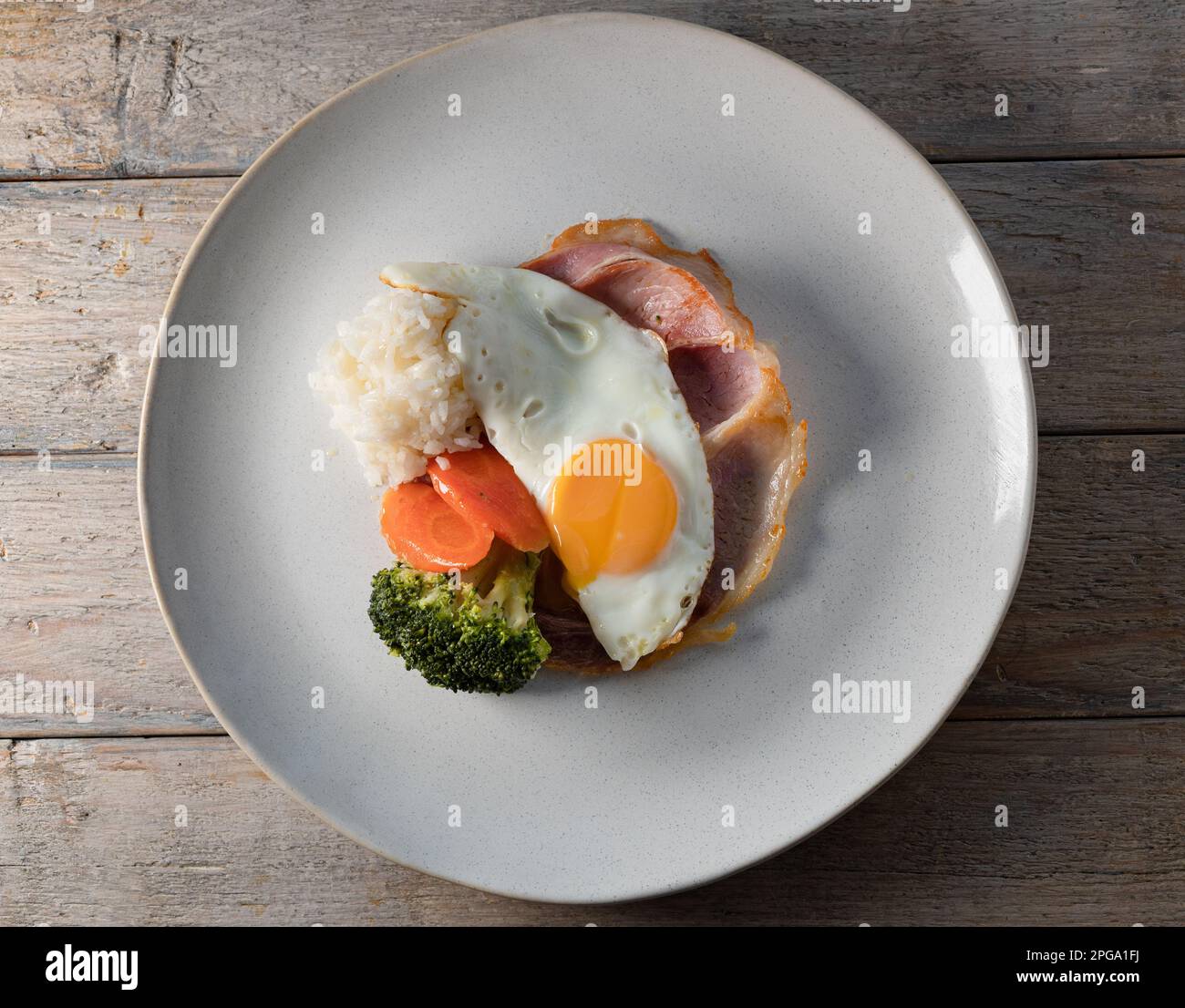 Eierfrühstück. Eier auf geröstetem Schinken mit Reis und gedämpftem Gemüse, Karotten und Brokkoli, serviert auf einem Teller auf einem Holztisch. Stockfoto