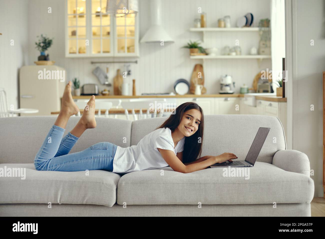 Süße 12s-jährige Frau benutzt Laptop auf dem Sofa im gemütlichen Wohnzimmer, posieren, lächeln, auf die Suchinformationen der Kamera schauen, Aufgaben machen, online lernen, mit chatten Stockfoto