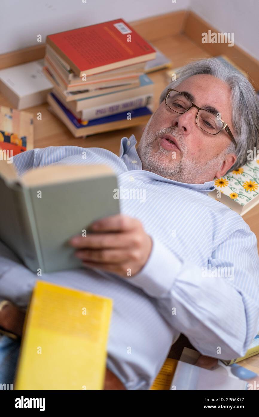 Der grauhaarige Mann, der bequem auf dem Boden liegt, liest faszinierend ein Interessantes Buch und ist überrascht, was geschrieben wird. Stockfoto