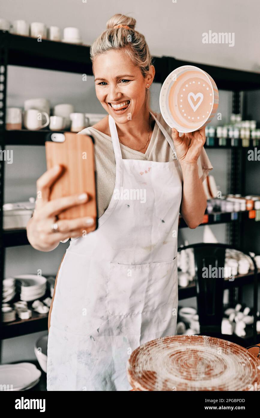 Ich bin so glücklich, wie das aussieht. Eine attraktive, reife Frau, die ihr Handy benutzt, um ein Selfie mit ihrer Töpferei in ihrem Studio zu machen. Stockfoto
