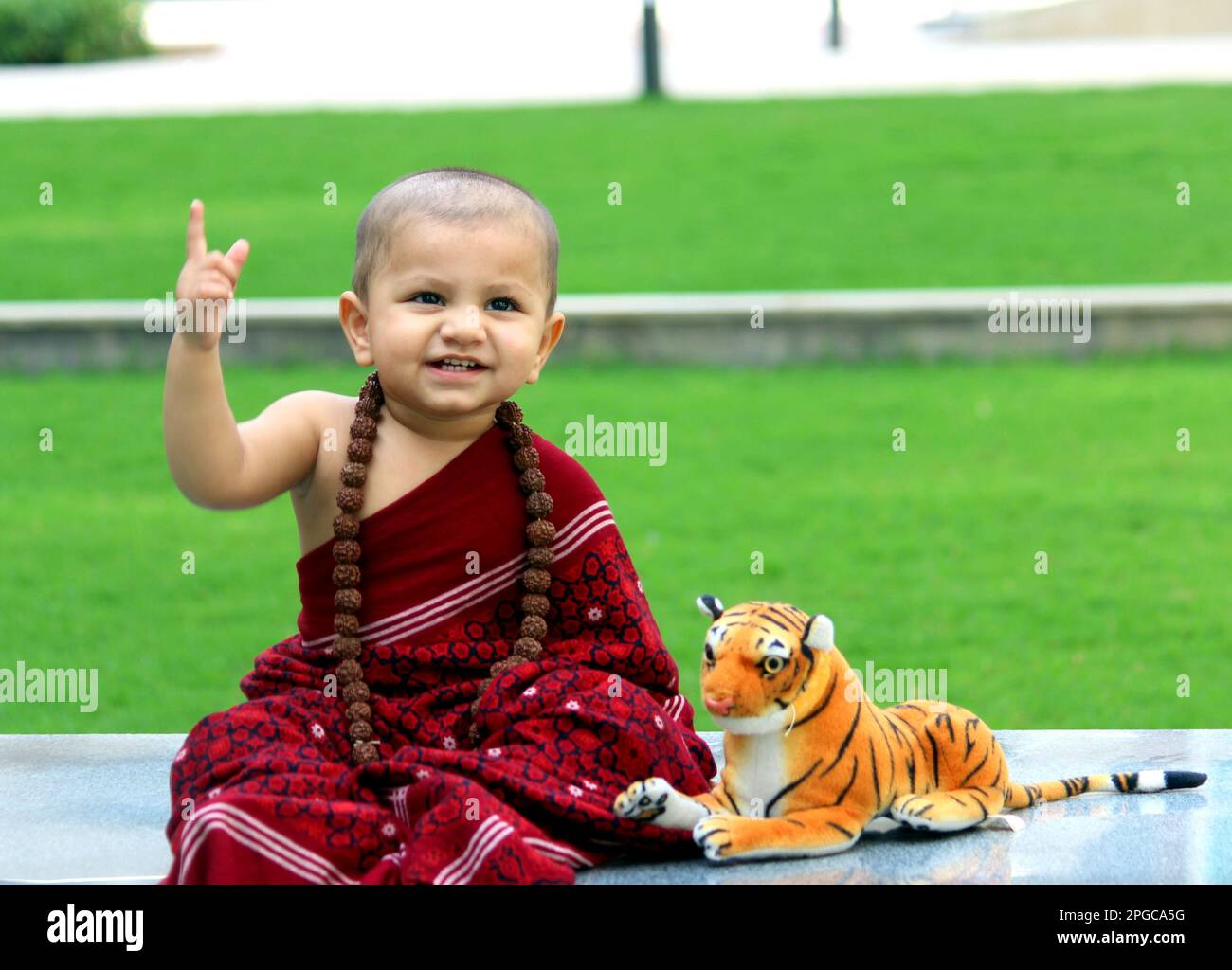 Ein süßer, kahlköpfiger Junge trägt Monk Avatar, einen kastanienbraunen Schal, Rudraksh, und ein süßes Lächeln. Ein Tiger-Spielzeug ist neben dem Jungen. Stockfoto