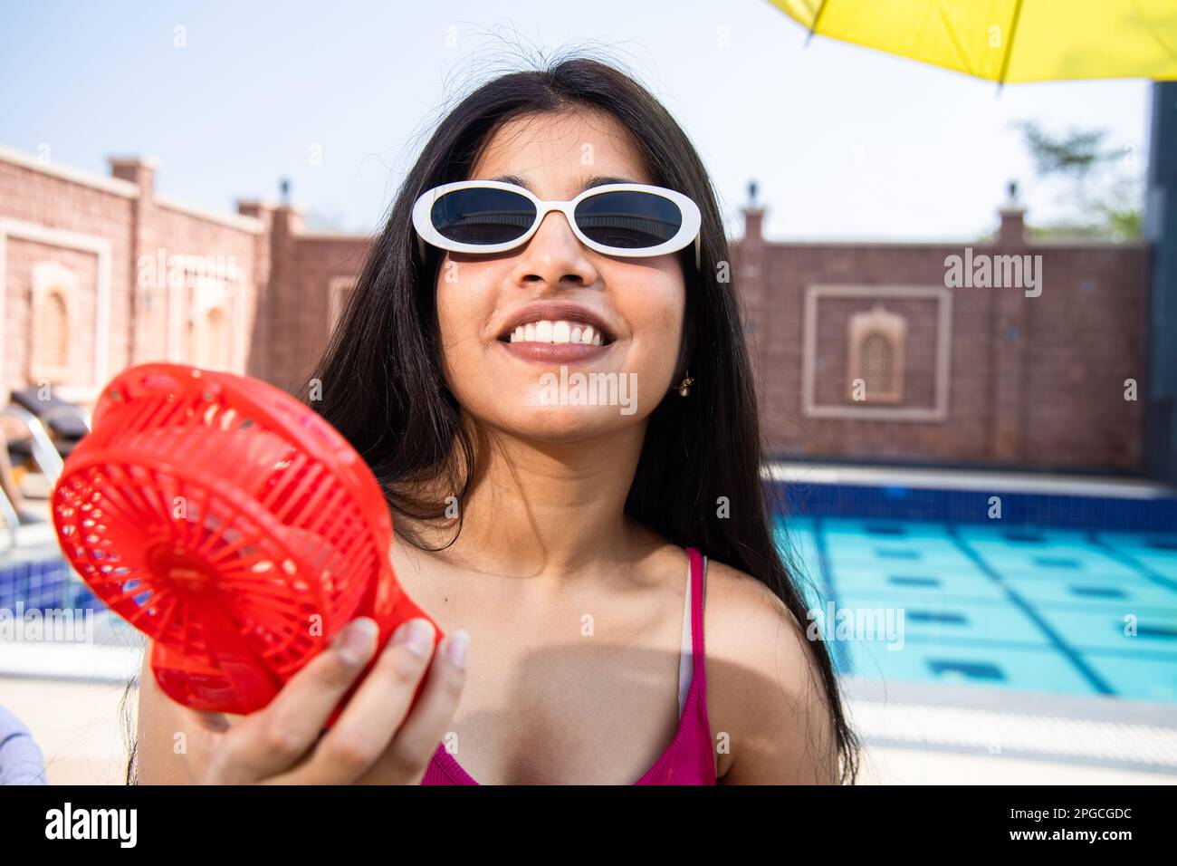 Junge indische Teenagerin, die eine Sonnenbrille trägt und den Luftstrom von einem tragbaren elektrischen Ventilator genießt und ihren Körper an sonnigen Tagen im Freien kühlt. Sommerhitze. Stockfoto
