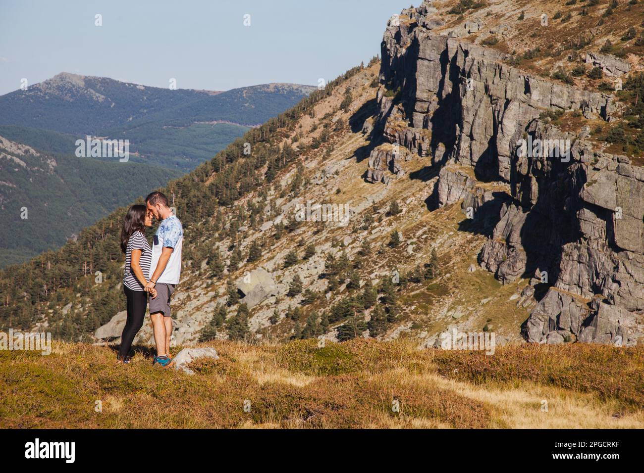 Seitenansicht eines liebevollen Paares in legerer Kleidung, das auf dem Rasen mit trockenem Gras steht und Händchen hält, während man den Sommerurlaub vor dem felsigen Berg i genießt Stockfoto
