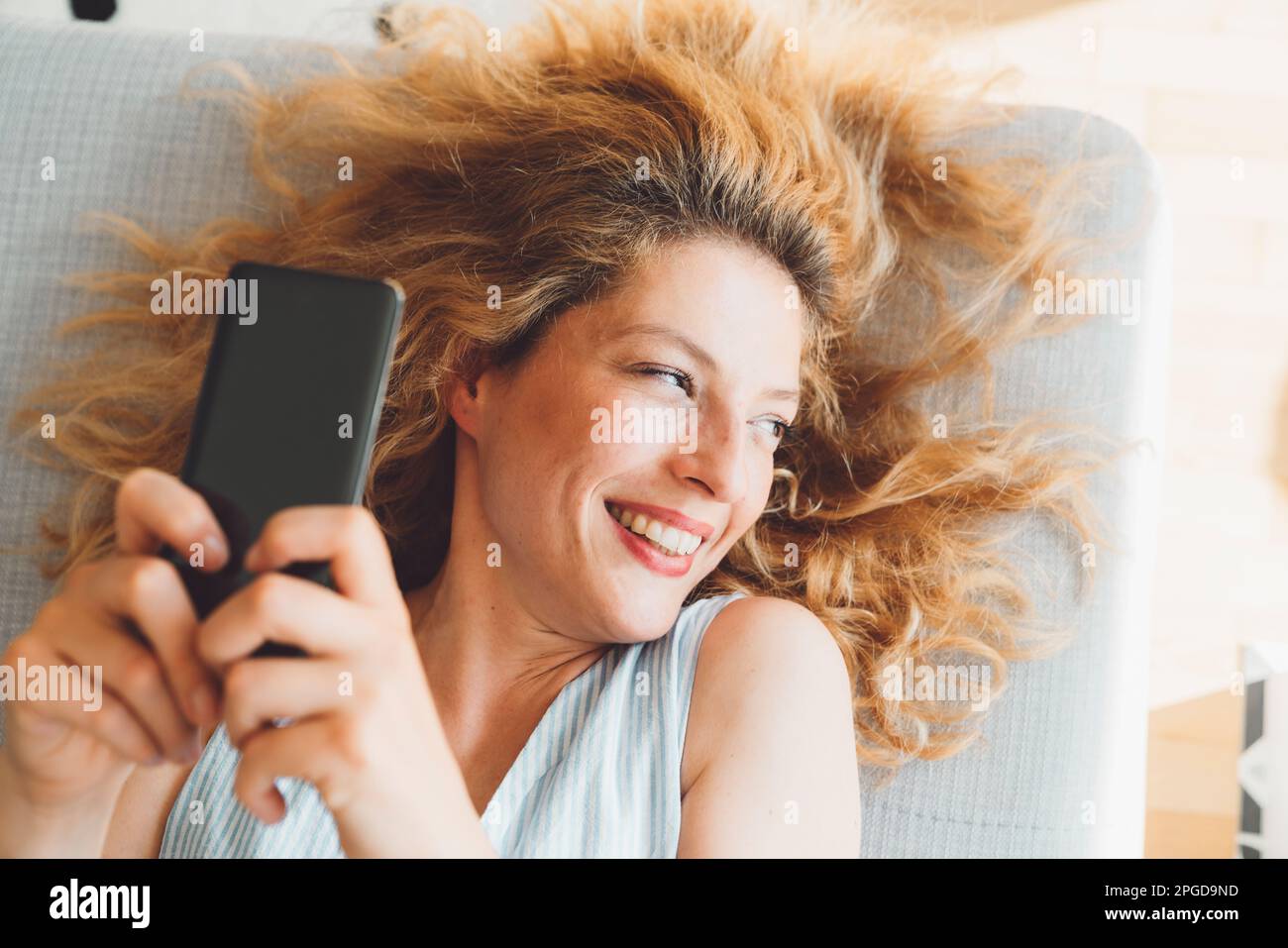 Nahaufnahme des Porträts einer Frau mit lockigem Haar, die wegschaut und auf der Couch liegt, während sie ihr Telefon hochhält und Selfies macht Stockfoto