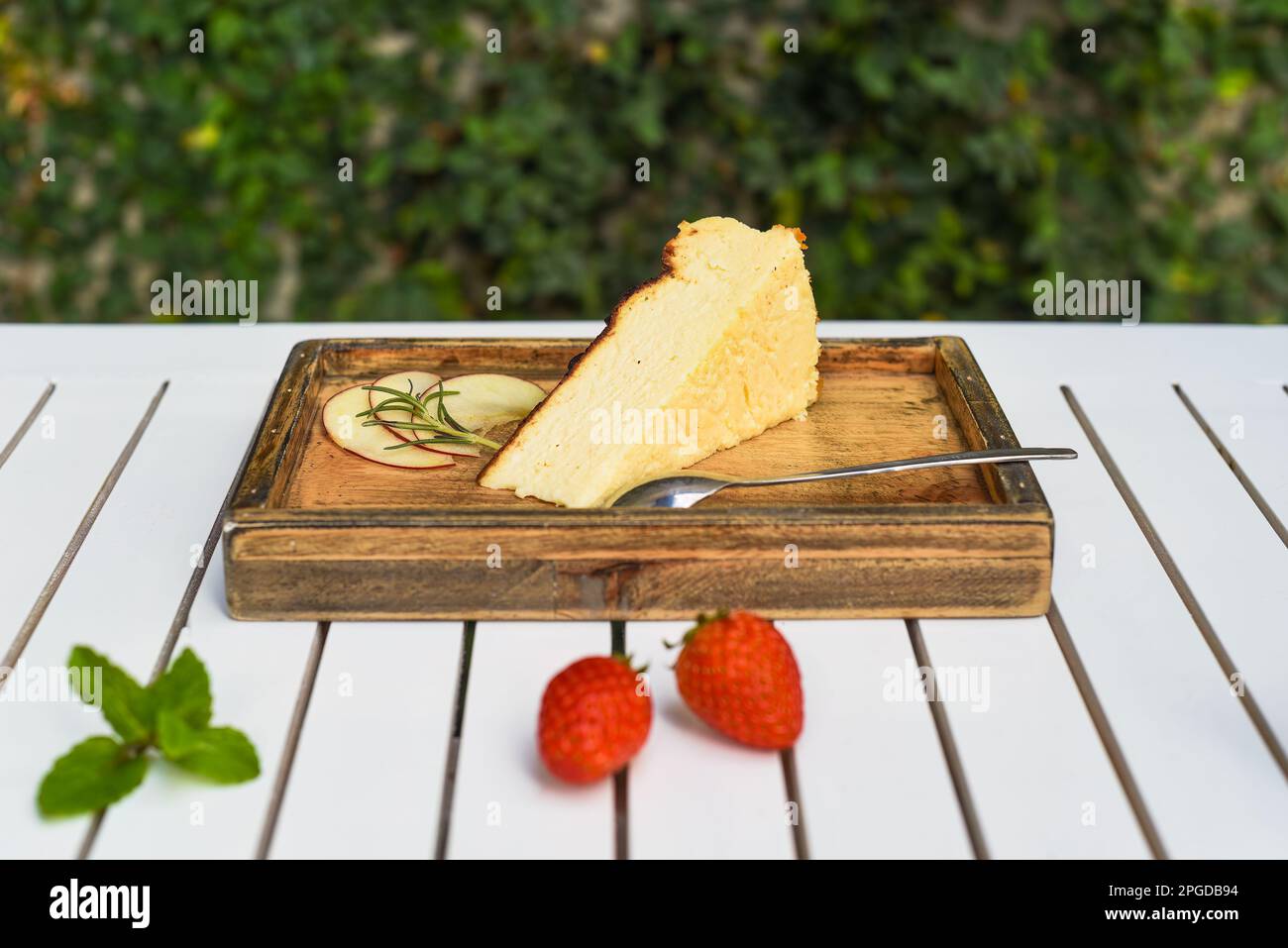 Ein Stück Käsekuchen auf einem Holzbrett mit Erdbeere und Minze auf einem weißen Holztisch mit grünen Blättern und einer Kopierfläche Stockfoto
