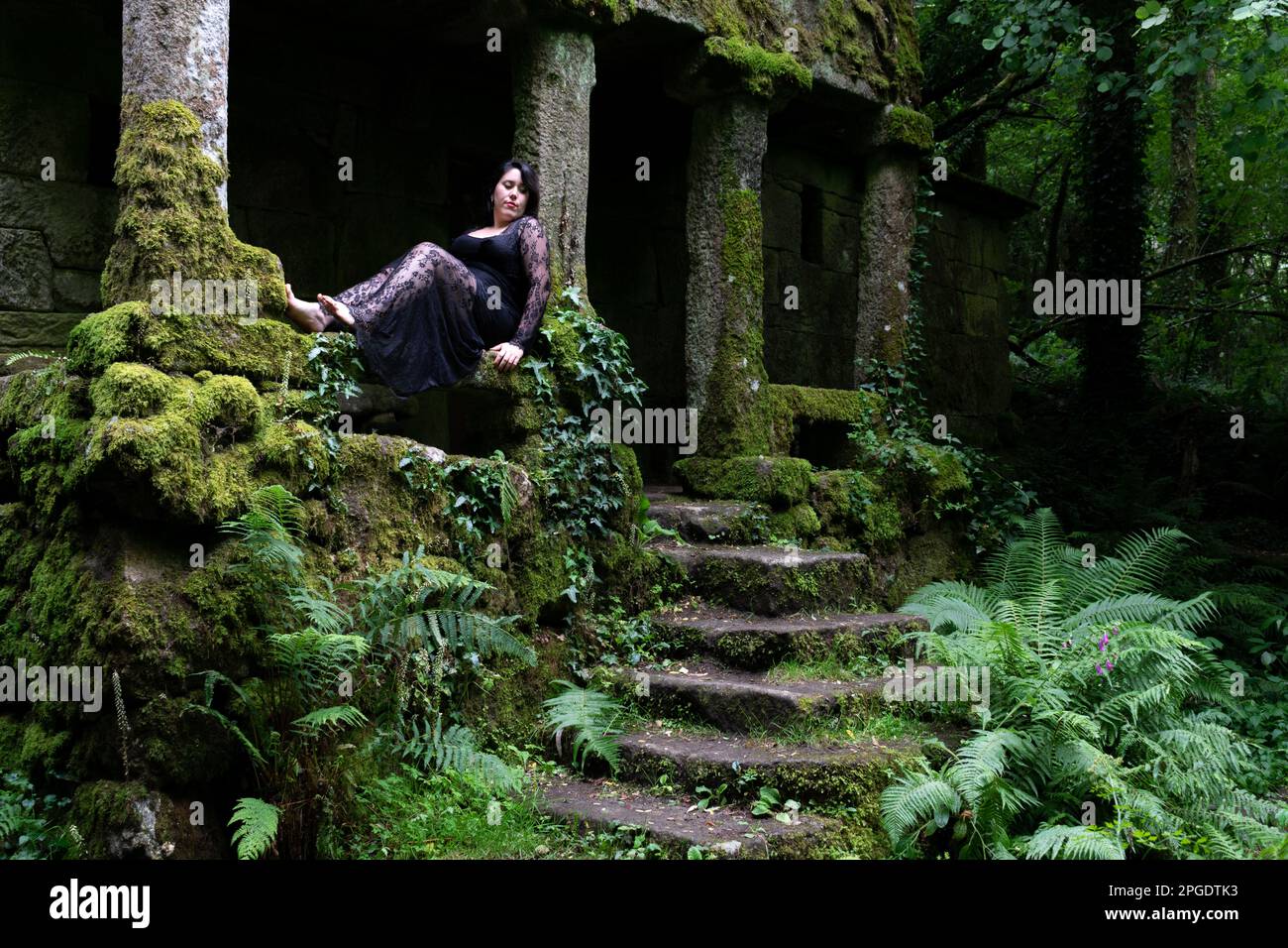 Eine Frau in einem langen schwarzen Kleid, die vor einem verlassenen Haus im Wald in Spanien sitzt Stockfoto