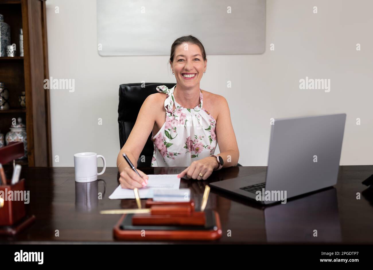 Eine glückliche Frau sitzt am Schreibtisch und schaut in die Kamera, während ihr Laptop vor ihr offen ist. Sie nutzt drahtlose Technologie für die Geschäftskommunikation Stockfoto