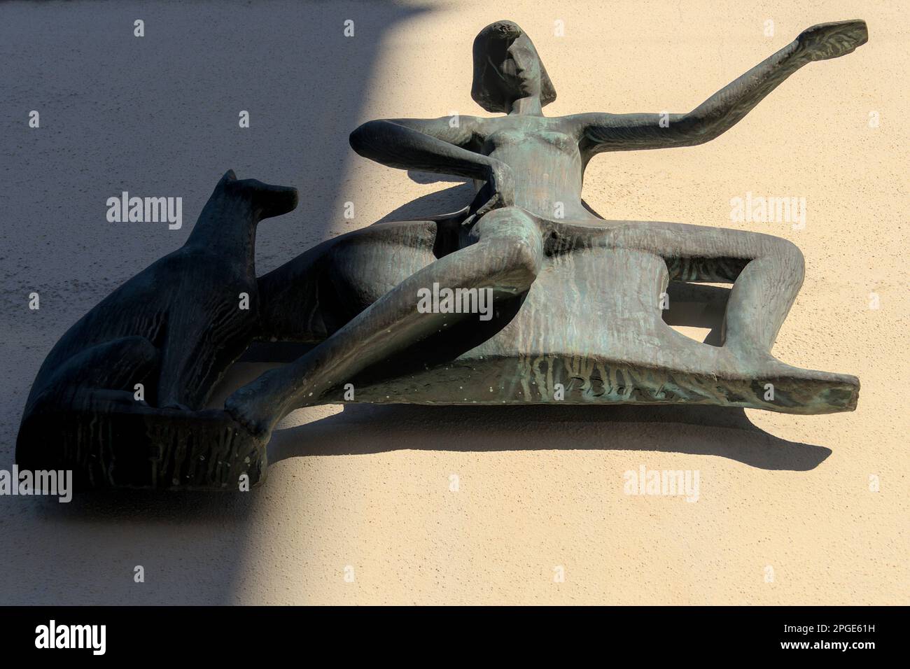 Abstrakte Bronzeskulptur, die eine Frau mit einem ausgestreckten Arm und einem Hund neben ihr zeigt, gekennzeichnet durch glatte, fließende Linien und eine grünliche Patina. Stockfoto