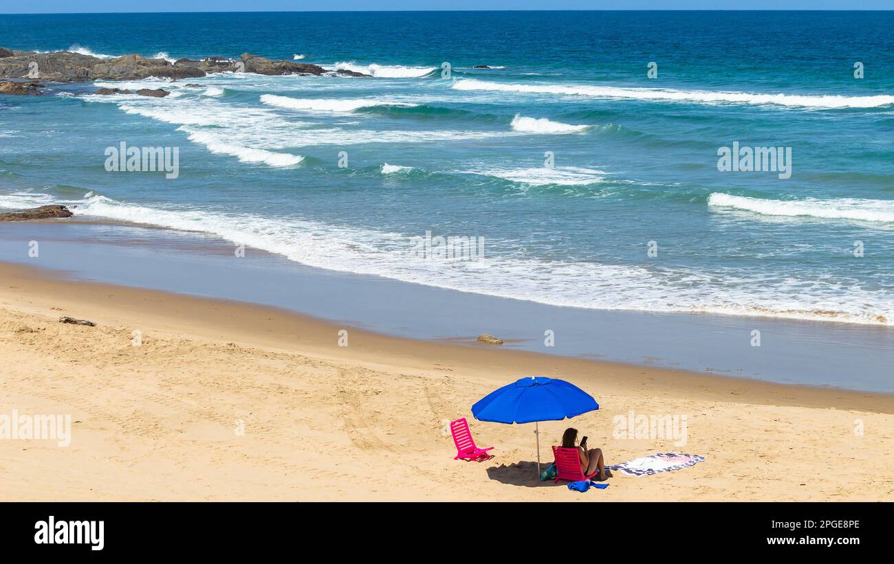 Sonniger Tag am Strand Weißes Mädchen, das auf pinkfarbenen Stühlen unter einem Sonnenschirm sitzt und sich mit dem mobilen blauen Wellenwasser des Ozeans an einer malerischen Urlaubslandschaft erholt. Stockfoto