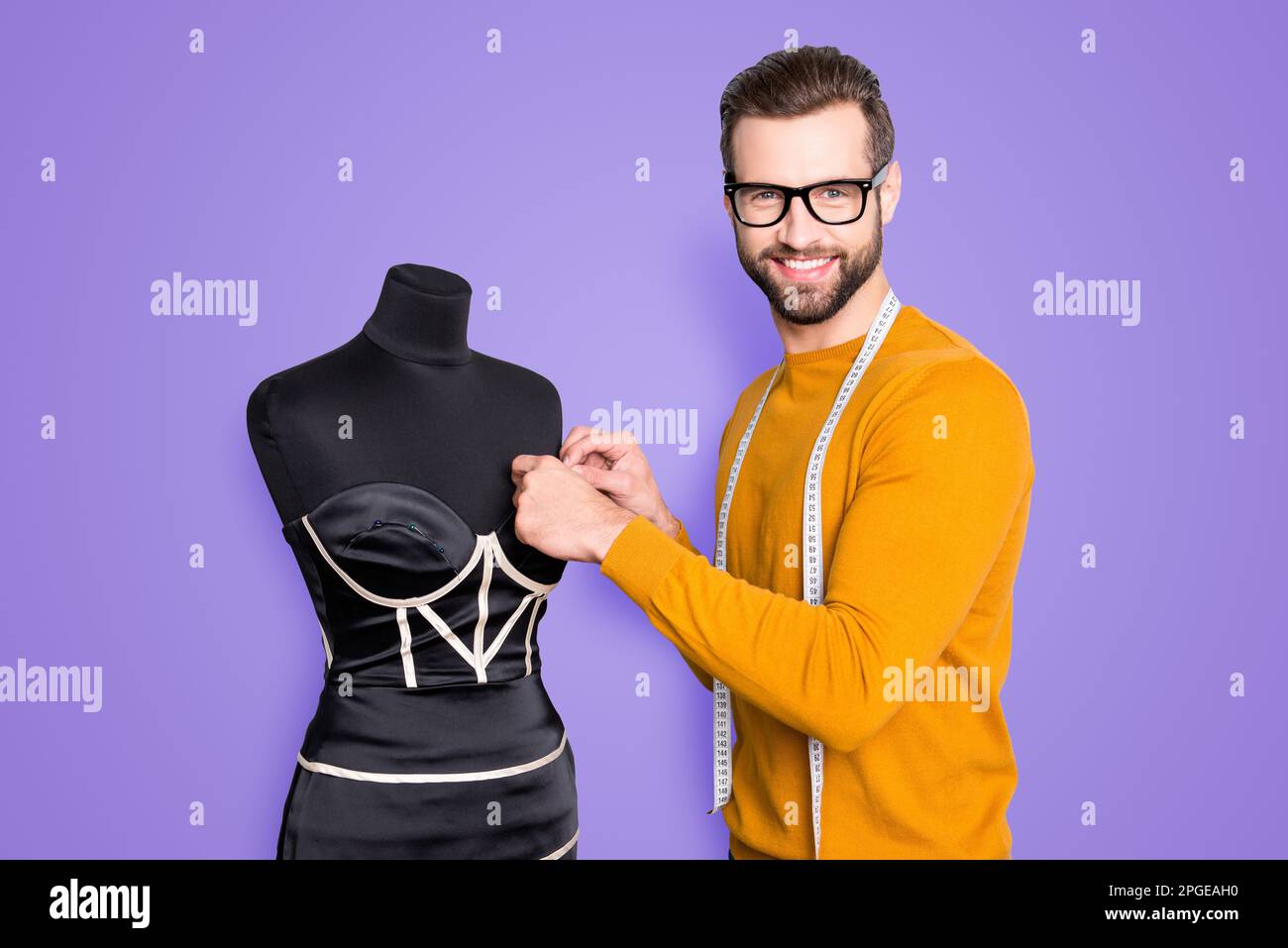 Portrait eines fröhlichen berühmten Schneiders im Pullover mit Zentimeter am Hals, der Messungen von eleganten Kleidern an Schaufensterpuppe mit Blick auf die Kamera durchführt Stockfoto