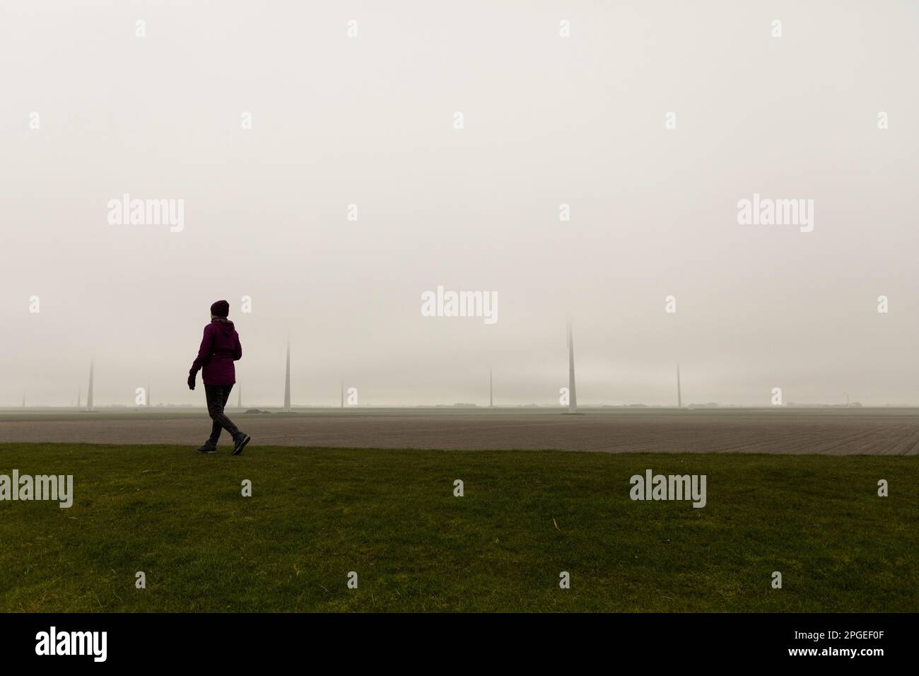 Eine Frau geht auf einem Deich in der Nähe von Eemshaven in Nordholland, während der Nebel Windturbinen im Hintergrund verschluckt Stockfoto